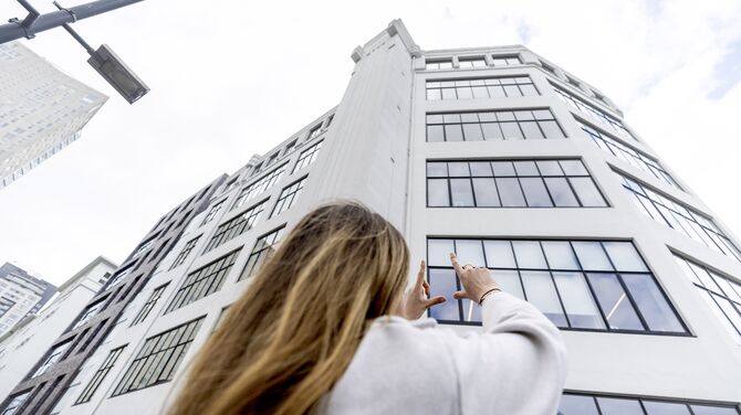 Femeie care își ridică mâinile spre o clădire albă de înălțime medie, cu multe ferestre.