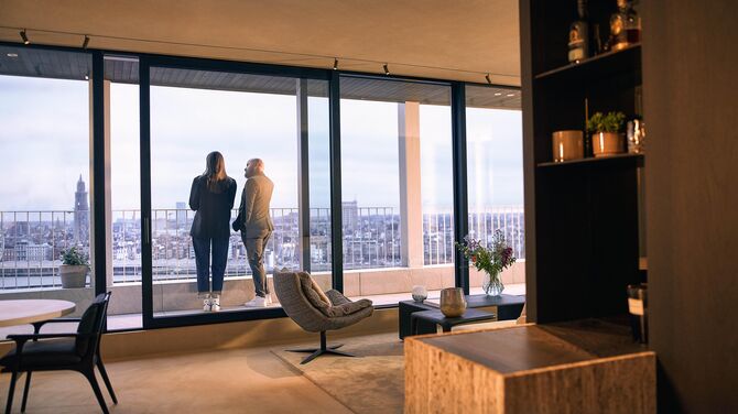 Alt: Vedere interioară a unui apartament modern cu uși glisante largi și două persoane pe un balcon.